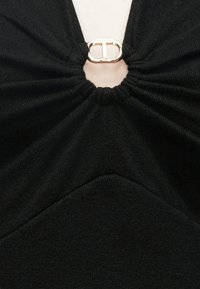 Sort stoftop med et tekstureret design og samlet stof ved halsudskæringen, med en guldfarvet hardwaredetalje i midten.