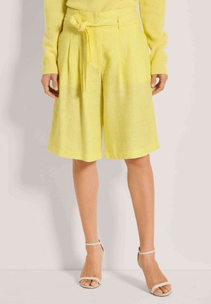 Culottes en lin jaune avec ceinture à nouer, dotées de plis, d'une coupe ample et d'une surface légèrement texturée ; portées avec des sandales blanches à brides.