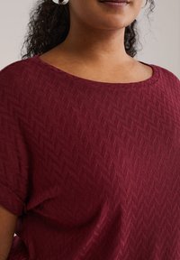 Bordeauxrotes, strukturiertes Oberteil mit Diamantmuster, rundem Ausschnitt und kurzen Ärmeln, aus leichtem Stoff gefertigt.