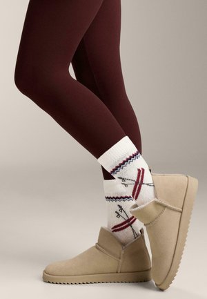 Bézs színű szarvasbőr bokacipők, lekerekített orral, alacsony sarokkal és oldalsó gombbal, fehér zoknival párosítva, amely piros és kék sí témájú mintákkal díszített.