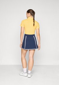 Camisa polo amarilla de canalé con mangas cortas, combinada con una falda azul marina con ribetes blancos. Zapatillas blancas y calcetines tobilleros completan el conjunto.