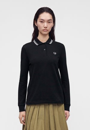 Vrouw met lang donker haar, gekleed in een zwarte polo met lange mouwen met witte bies en een beige geplooide rok, staand tegen een effen achtergrond.