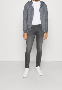 Sudadera gris con cremallera y bolsillo canguro, llevada sobre una camiseta blanca. Combinada con jeans ajustados gris oscuro y zapatillas blancas.