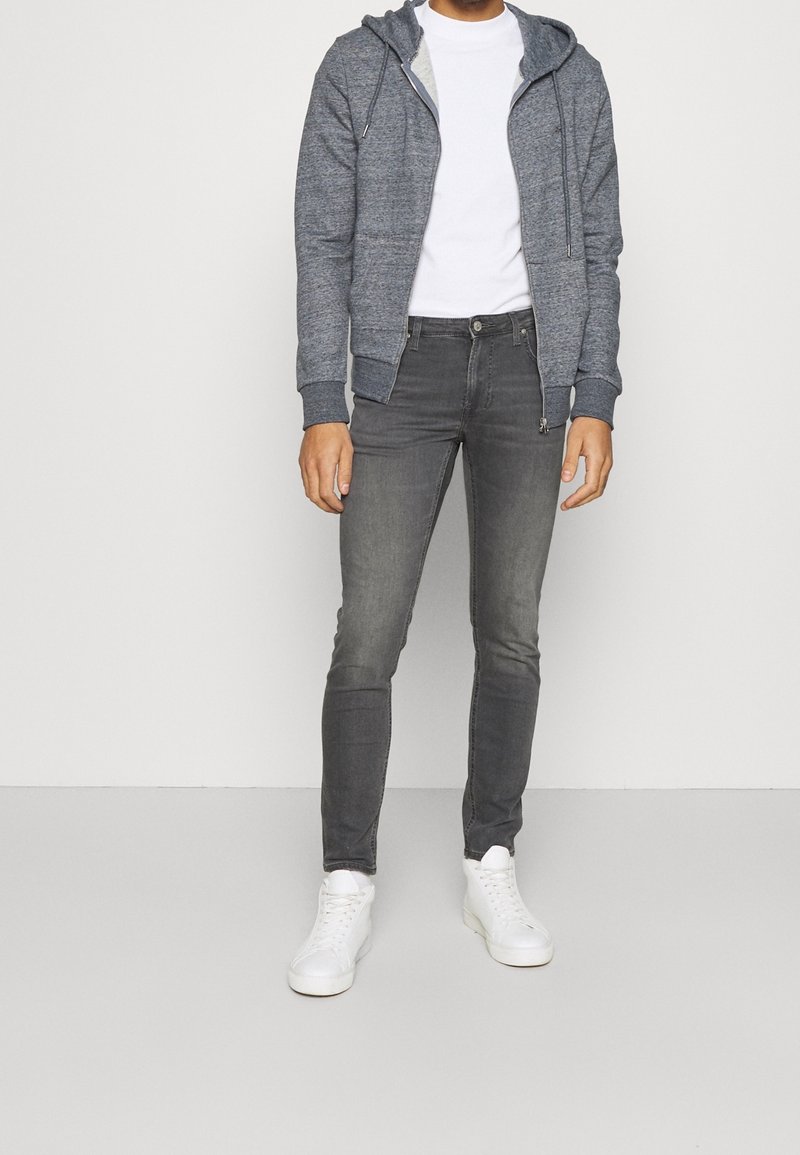 Sudadera gris con cremallera y bolsillo canguro, llevada sobre una camiseta blanca. Combinada con jeans ajustados gris oscuro y zapatillas blancas.