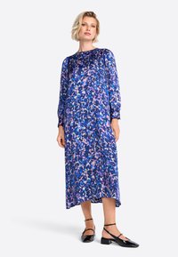 Langes, fließendes Kleid in einem abstrakten Muster aus Blau, Lila und Pink. Mit langen Ärmeln und einem runden Ausschnitt ausgestattet. Kombiniert mit schwarzen, flachen Schuhen.