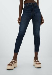 Jeans skinnys de tiro alto en azul oscuro, con costuras clásicas, bolsillos delanteros y una textura suave, combinados con sandalias de plataforma en color tostado.