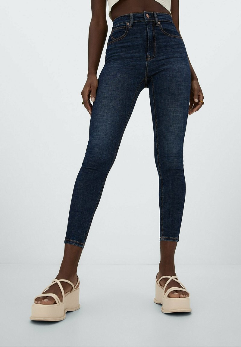 Jeans skinnys de tiro alto en azul oscuro, con costuras clásicas, bolsillos delanteros y una textura suave, combinados con sandalias de plataforma en color tostado.