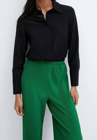 Chemise noire à manches longues avec col, associée à un pantalon large plissé vert doté d'une taille élastique. Tissu lisse et couleurs vives.