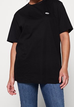 Personne portant un t-shirt noir ample à manches courtes avec un petit logo blanc sur la poitrine gauche et un jean en denim bleu.