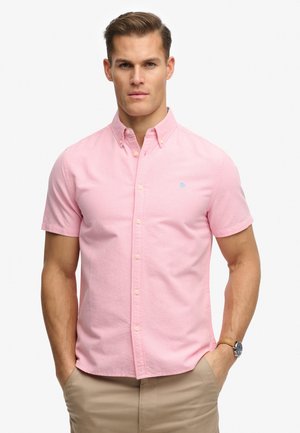 Hombre con camisa de manga corta color rosa claro abotonada y pantalones beige con las manos en los bolsillos, de pie sobre un fondo blanco.