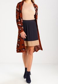 Cardigan lungo color marrone arancio con stampa leopardata nera, abbinato a un top beige a costine, una gonna plissettata blu navy e stivaletti blu navy.