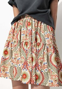 Jupe à volants florale en crème, ornée de motifs rouges, verts et bleus. Taille élastique avec une texture froncée, accentuant le volume et le mouvement.