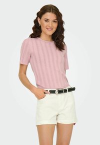 Top a maglia rosa chiaro con maniche corte e texture a coste, abbinato a pantaloni corti bianchi arrotolati e una cintura nera con borchie.