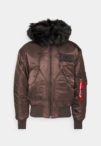 Hnědá bunda typu bomber s černou koženkovou kapucí, předními kapsami, žebrovanými manžetami a spodním lemem, a červeným štítkem "Odstranit před letem" na kapse na rukávu.