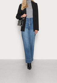 Blazer noir sur un col roulé gris, associé à un jean en denim bleu à jambes larges avec des boutons sur le devant, et des bottines noires.