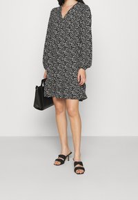 Valkoinen ja musta kuviollinen mekko, jossa on V-pääntie ja pallohihat, helma levenee. Yhdistetty mustiin korkokenkiin ja mustaan käsilaukkuun.