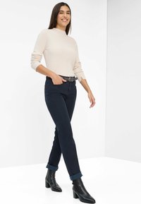 Pull en maille beige, jean ajusté bleu marine avec les poignets retroussés, et bottines à talons noires, sur fond blanc uni.