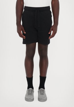 Schwarze Shorts aus weichem Material mit elastischem Bund und verstellbarem Kordelzug, ausgestattet mit zwei Fronttaschen. Kombiniert mit grauen Sneakers.