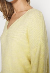 Gelber Strickpullover mit V-Ausschnitt, geripptem Muster und weiten Ärmeln für eine lockere Passform. Weicher Stoff mit einem subtilen Glanz.