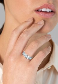 Silberner Ring mit einer Reihe von fünf klaren Edelsteinen, glatte Oberfläche und abgerundete Kanten, getragen an einer Hand in der Nähe eines Gesichts.
