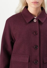Manteau en laine bordeaux avec un col évasé, cinq boutons et deux poches latérales. Texture douce avec une coupe structurée.