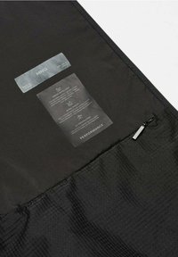 Tissu noir avec étiquettes de soin et de performance cousues, détaillant la déperlance à l'eau, la protection contre le vent, la protection UV et une fermeture éclair visible sur un matériau texturé.