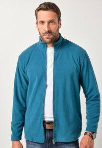 Giacca in pile turchese con colletto alto, zip frontale, e maniche lunghe; abbinata a una maglietta bianca e jeans in denim.