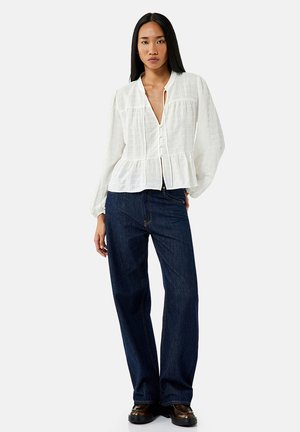 Witte, lichte button-up blouse met lange mouwen, gerimpelde peplum taille en subtiele kreukelstructuur; gecombineerd met donkerblauwe wijde jeans.