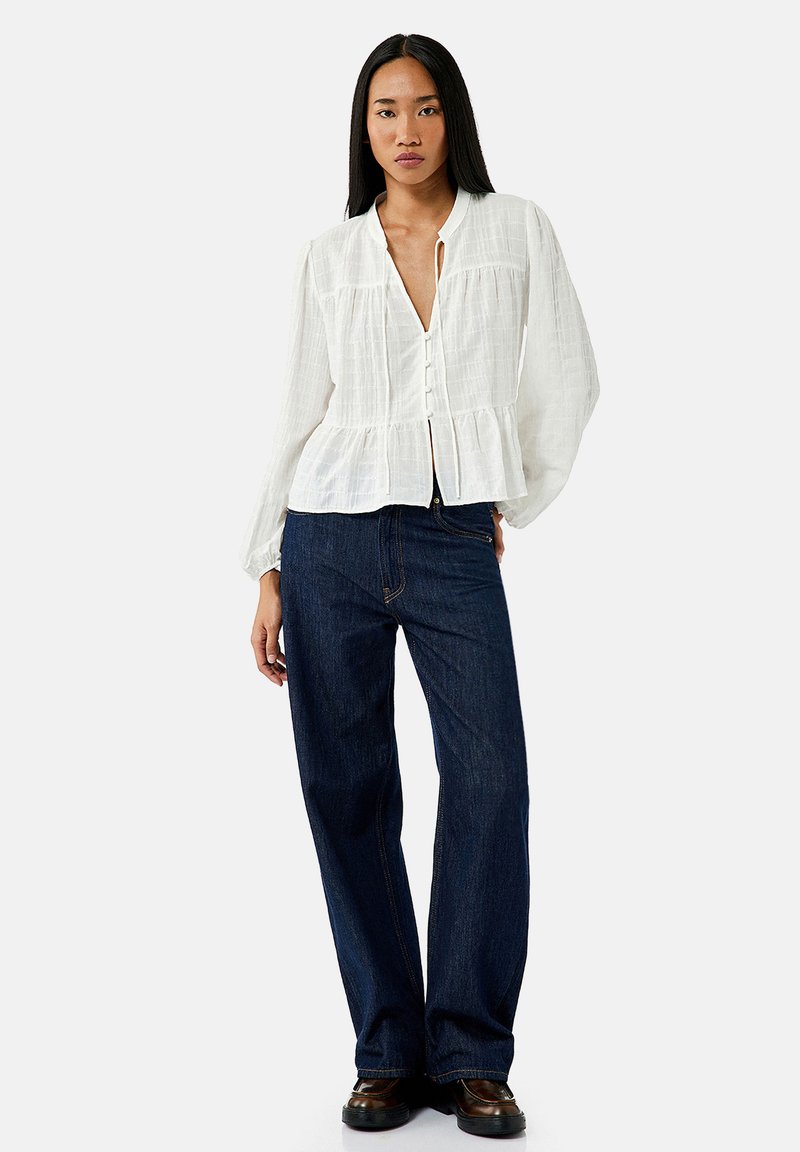 Witte, lichte button-up blouse met lange mouwen, gerimpelde peplum taille en subtiele kreukelstructuur; gecombineerd met donkerblauwe wijde jeans.