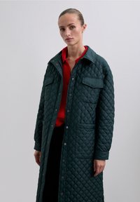 Abrigo largo acolchado en verde oscuro, con cuello, dos bolsillos frontales y cierre de botones. Lleva puesto sobre una blusa roja. Textura suave.