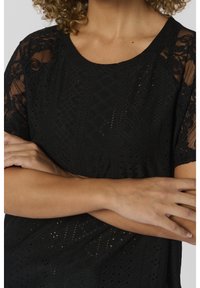 Svart blus med ett öppet mönster med diamantformer, korta genomskinliga ärmar och rund halsringning. Mjuk textur och avslappnad passform.