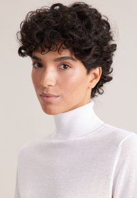 Biały sweter typu golf wykonany z materiału o drobnym ściegu, z wysokim kołnierzem i bezszwowym designem, noszony przez osobę z kręconymi włosami.