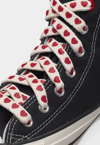 Μαύρο αθλητικό παπούτσι με λευκά κορδόνια που φέρουν κόκκινα καρδιάκια, περασμένα από μεταλλικά κουμπώματα, με ορατή λευκή ραφή στο παπούτσι.