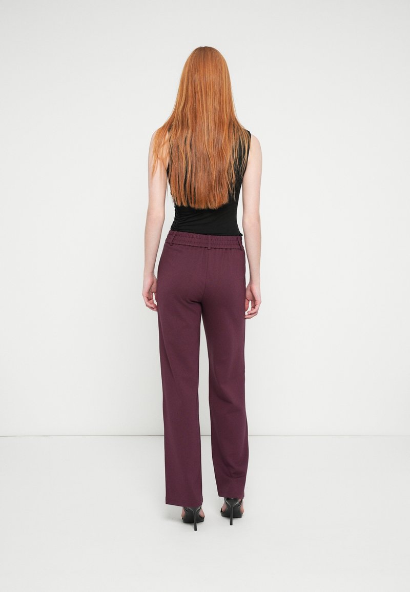 Femme aux longs cheveux roux, debout face à un mur blanc, portant un haut noir sans manches, un pantalon violet et des chaussures à talons noirs.