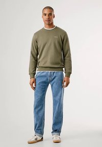 Sudadera verde oliva con puños y dobladillo acanalados, combinada con vaqueros azul claro y zapatillas blancas con detalles en negro.