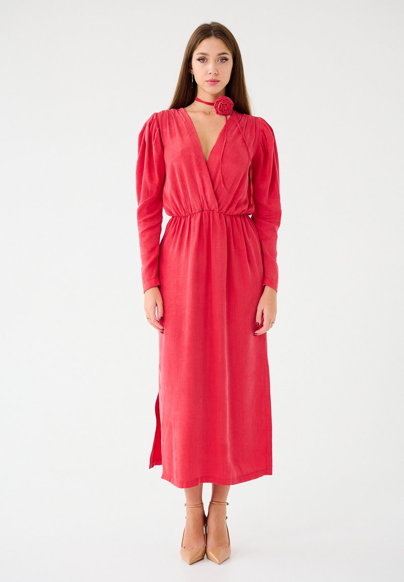 Robe rouge à manches longues croisée avec des épaules bouffantes, une taille cintrée et une fente sur le côté. Caractérisée par un décolleté en V profond et une accent floral au niveau du cou.