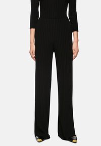 Black ribbed trousers with a high waist, wide legs, and textured fabric. Paired with black shoes featuring a gold accent on the toe.