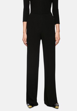 Black ribbed trousers with a high waist, wide legs, and textured fabric. Paired with black shoes featuring a gold accent on the toe.