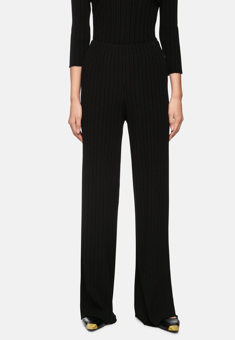 Black ribbed trousers with a high waist, wide legs, and textured fabric. Paired with black shoes featuring a gold accent on the toe.