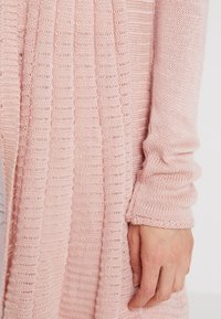 Ljusrosa stickad kofta med ribbad textur, långa ärmar och en lös passform. Tyget ser mjukt och lätt ut.