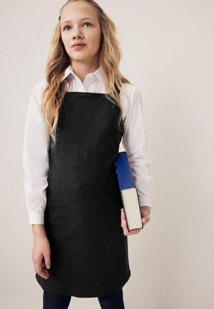 Giovane donna con lunghi capelli biondi, grembiule nero e camicia bianca, che tiene un libro spesso blu e bianco su uno sfondo semplice.