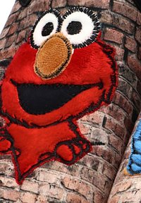 Figura de peluche roja de un personaje de dibujos animados con ojos grandes y una cara bordada, contra un fondo de ladrillo gris texturizado.