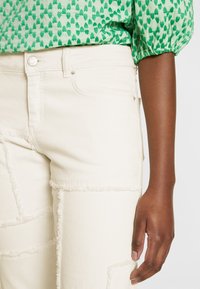 Personne portant un jean blanc taille haute avec des détails effilochés et un chemisier vert à motifs avec des manches bouffantes.
