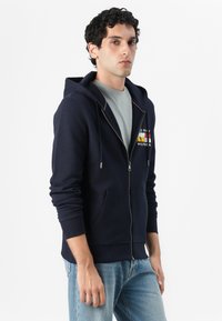 Sweat-shirt à zip bleu marine en mélange de coton, avec une poche avant, un capuchon à cordon de serrage et un design de logo multicolore sur la poitrine.