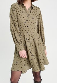 Robe boutonnie couleur vert olive avec des motifs floraux noirs, manches longues et jupe plissée. Tissu lisse avec une coupe décontractée.