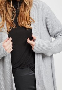 Femme portant un cardigan en maille côtelée gris clair par-dessus un haut noir et un pantalon en cuir noir, ajustant les bords avant du cardigan.