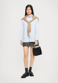 Donna che indossa una camicia azzurro chiaro con un maglione beige sulle spalle, una gonna mini nera, calze scure con mocassini e tiene una borsa nera.