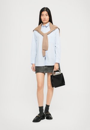 Donna che indossa una camicia azzurro chiaro con un maglione beige sulle spalle, una gonna mini nera, calze scure con mocassini e tiene una borsa nera.