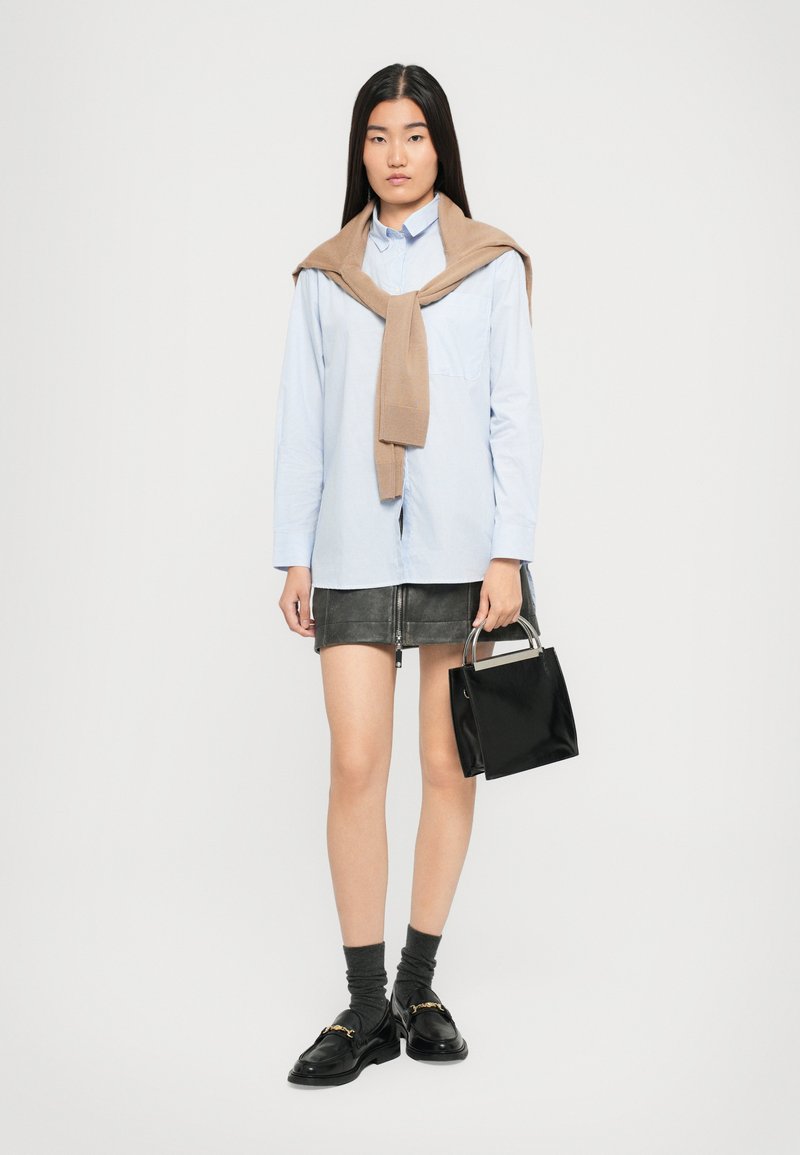 Frau in hellblauem Shirt mit beigem Pullover über den Schultern, schwarzer Minirock, dunklen Socken mit Loafers und einer schwarzen Handtasche.
