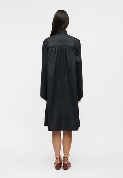 Femme debout de dos, portant une robe noire à manches longues de longueur moyenne et des chaussures plates marron à lacets sur fond blanc.